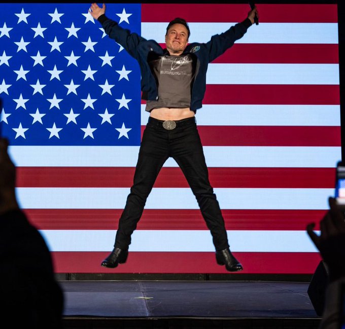 A man in a dark jacket and t-shirt jumping in mid-air with arms raised in front of a large American flag backdrop.