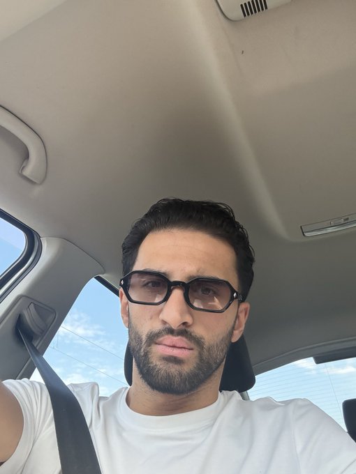 A selfie of a young man with dark hair, a beard, and black sunglasses wearing a white t-shirt while sitting in a car with a seatbelt on.