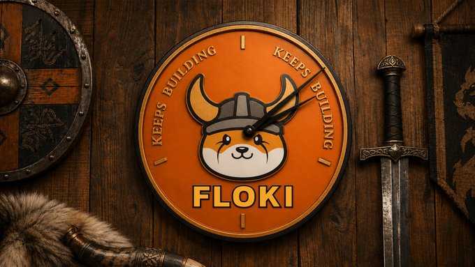 A Viking-themed wall clock featuring a Shiba Inu dog wearing a horned helmet is displayed with "FLOKI" written at the bottom, surrounded by the phrase "KEEPS BUILDING" repeated around its face. The clock is set against a rustic wooden background alongside a medieval sword and shield, symbolizing the "24/7" continuous building effort of the FLOKI cryptocurrency project.