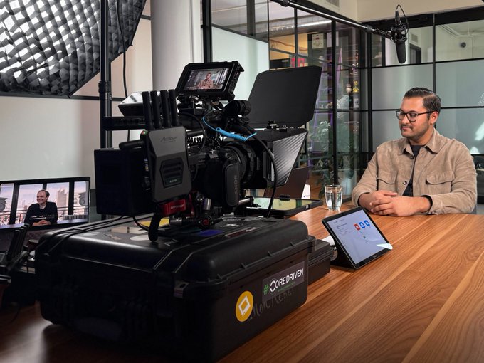 A professional video production setup with a large camera rig sits on a wooden desk, where a man in a beige jacket sits across from a tablet during what appears to be a filmed interview, with branding reading "COREDRIVEN" and "MOSSEN" visible on the equipment case. The scene captures the behind-the-scenes reality of creating content—literally embodying the idea that "life is a movie" by showing the filmmaking apparatus that transforms an everyday conversation into a polished production.