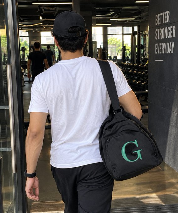 A man viewed from behind wearing a black cap, white t-shirt, and black pants, carrying a black Gymshark gym bag with a green 'G' logo, walking into a gym with 'BETTER STRONGER EVERYDAY' written on the wall.