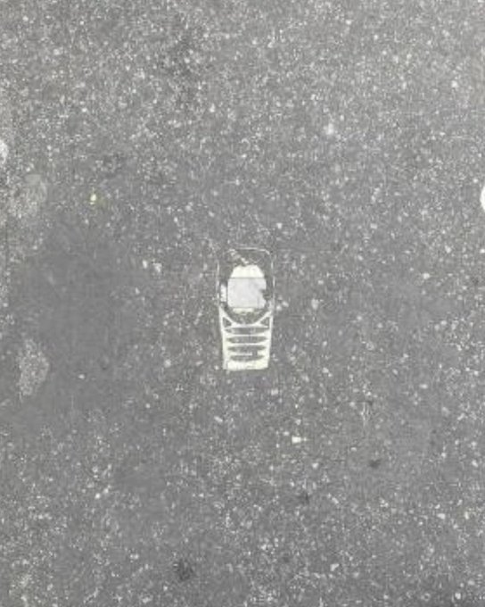 A small, rectangular silver object, possibly a piece of jewelry or a charm, rests on a textured gray asphalt surface.