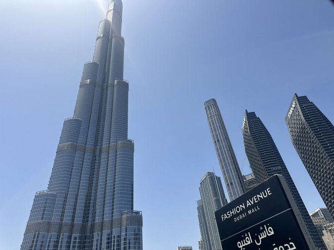 The image shows a tall building, likely the Burj Khalifa, which is an iconic skyscraper located in Dubai, United Arab Emirates. The photo captures the perspective of looking up at the building from below, emphasizing its height and grandeur. The surrounding cityscape features several other buildings, indicating that this location is situated within a bustling urban environment.