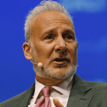 A close-up of an older man with gray hair and a beard wearing a suit and pink tie, speaking into a small microphone against a blue background.