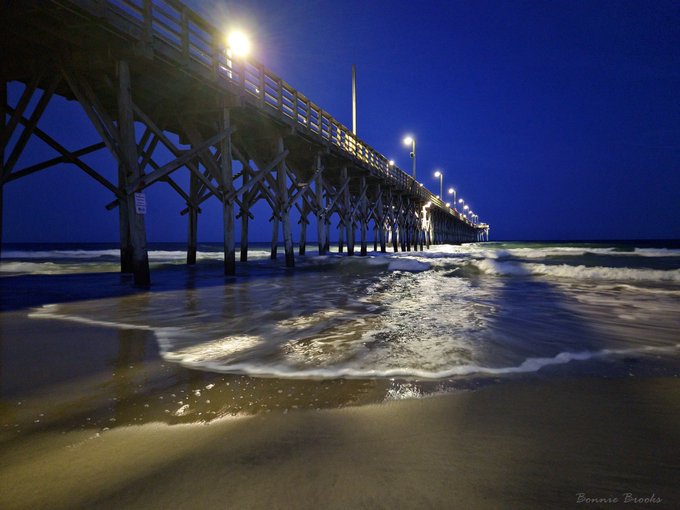 A nighttime photograph of a long wooden pier illuminated by lights, with waves washing onto the sandy beach in the foreground, credited to Bonnie Brooks.