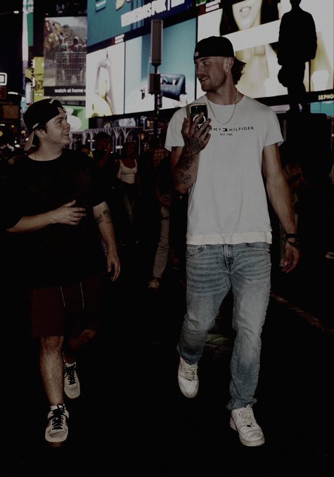 Two men walking through a neon-lit city street at night, one in a black shirt and shorts looking up, the other taller man in a Tommy Hilfiger shirt looking at his phone.