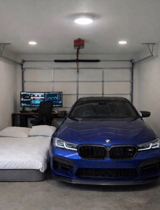 A blue BMW sedan is parked inside a garage that doubles as a bedroom, with a neatly made bed and a desk featuring a triple-monitor gaming/work setup just feet away from the car. The image embodies the post's sentiment of having all essentials in one space: a high-performance vehicle, a comfortable place to sleep, and an entertainment station.