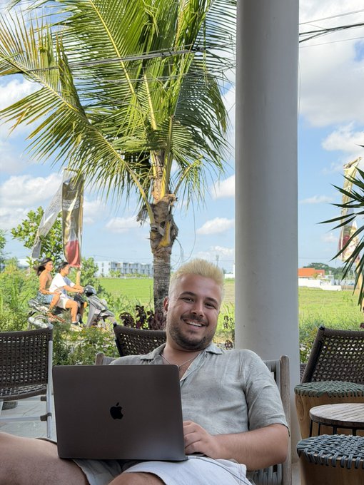A man with blonde hair smiles while working on his Apple MacBook at an outdoor café, surrounded by tropical palm trees and rice fields in the background. The image reflects the post's message of balancing trading with enjoying life's beautiful moments, embodying the "live for trading, trade for life" philosophy.