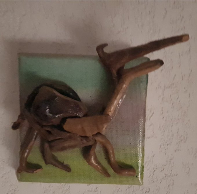 A small, abstract bronze-colored sculpture with spiky limbs rests on a greenish square base mounted against a textured white wall.