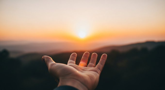 A hand is held open towards a bright sun setting behind distant mountains in a warm, hazy landscape.