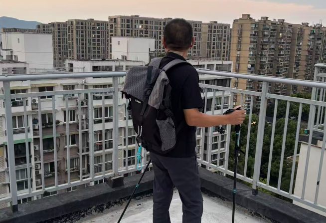 A man with a grey backpack stands on a balcony overlooking a dense cityscape of high-rise apartment buildings, holding a collapsed tripod.