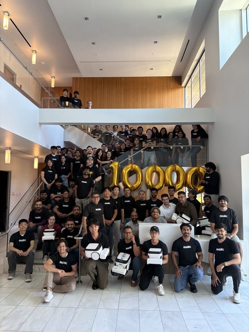 A large group of team members wearing matching black "matic" t-shirts poses on a modern staircase, holding white box packages with giant gold "10000" balloons prominently displayed in the center, celebrating the milestone of shipping their 10,000th product.