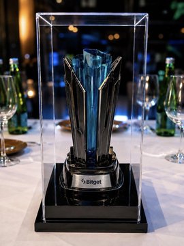 A sleek trophy encased in a clear display box with a teal centerpiece, featuring a plaque that reads "5tiget," sits on a white table flanked by wine glasses, likely representing one of the awards given to the UEX Global Alpha Tournament winners.