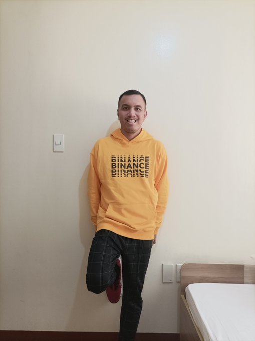 The image features a man standing against a white wall, smiling for the camera while wearing a yellow hoodie with black text that says "BINANCE". The background of the photo includes a bed and a chair, suggesting that this scene takes place in a bedroom or living space. The caption to the image reads: "Celebrating the re-entry of @binance in the Philippines by wearing this swag!".
