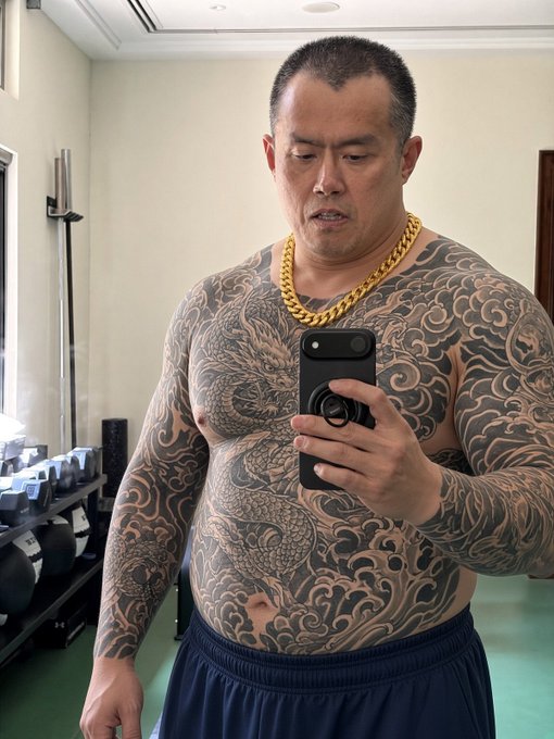 A heavily tattooed, muscular man wearing a thick gold chain takes a mirror selfie with a smartphone in what appears to be a gym setting.