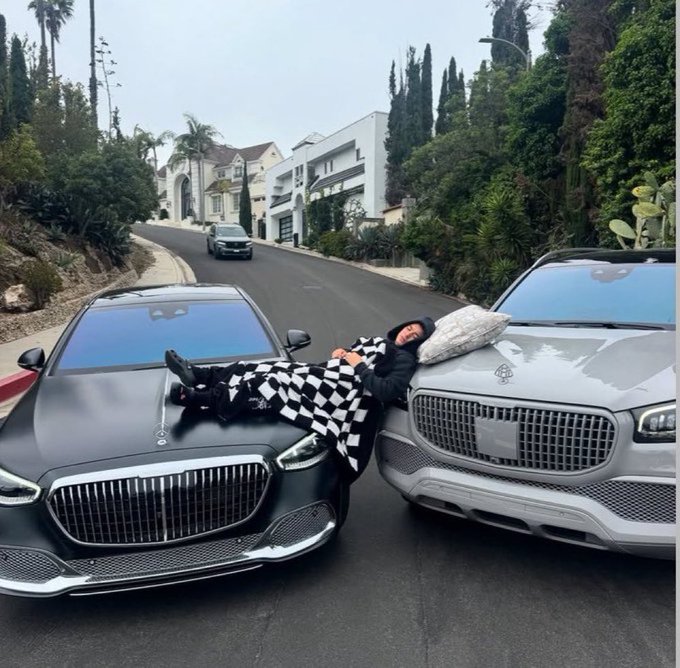A person wearing a black-and-white checkered outfit reclining between a black and a white luxury SUV parked on a residential street.