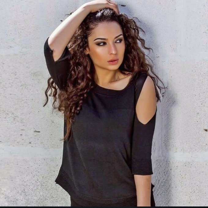 A model with long curly brown hair poses against a textured wall, wearing a black sweater with shoulder cutouts.