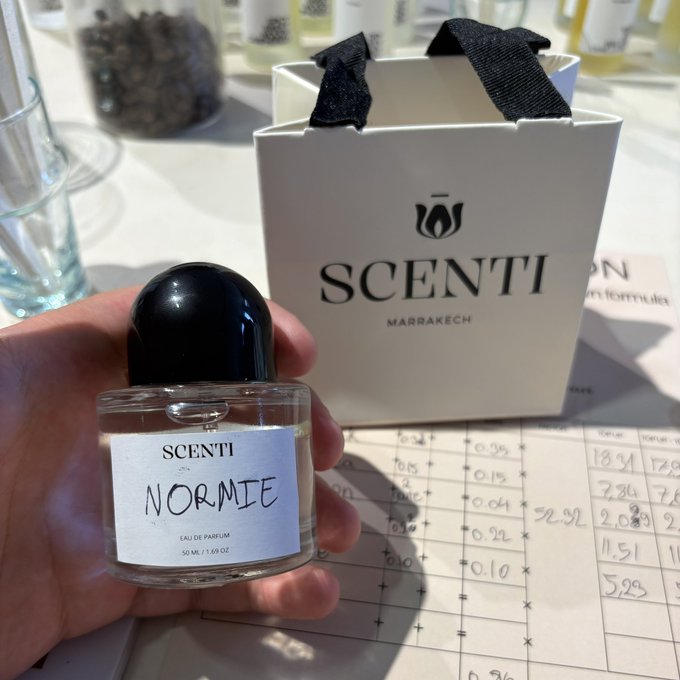A hand holds a small bottle of perfume labeled 'SCENTI NORMIE' in front of a white 'SCENTI MARRAKECH' gift bag and a handwritten fragrance formulation chart.
