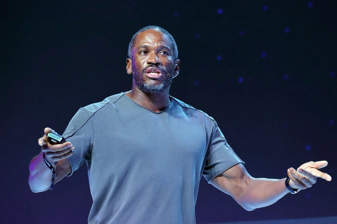 Actor Boris Kodjoe wearing a dark gray t-shirt, gesturing with both hands while holding a small clicker device during a presentation on a dark stage.