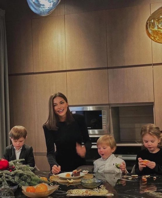 A woman stands smiling with three children at a table of food in a modern kitchen, with decorative greenery and fruit in the foreground.