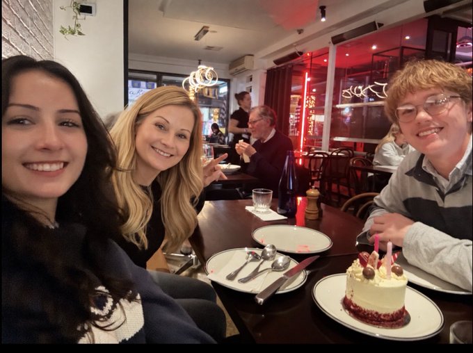 Three people smiling at a restaurant table with a birthday cake decorated with cherries and candles.