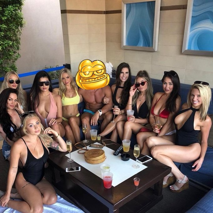 A group of people in swimwear posing together at what appears to be a pool party or beach club, with one person's face covered by a smirking emoji; drinks and a round container sit on the table in front of them.