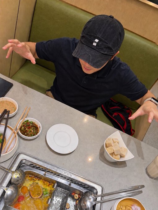 A person wearing a black cap and dark polo shirt sits at a restaurant table with hot pot food, sauces, and dipping ingredients, reaching their hand forward toward the camera.