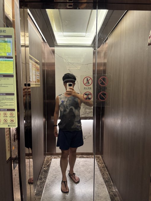 A person takes a mirror selfie inside an elevator wearing a cap, sleeveless shirt, and shorts while holding a phone.