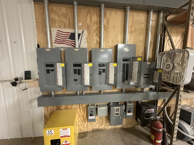 A row of heavy-duty electrical breaker panels and disconnects mounted on a plywood wall, with an OSHA safety cabinet and fire extinguisher below, supporting the post's claim of an 800-amp industrial-scale electrical setup capable of powering approximately 20,000 phones plus additional ASIC mining hardware.
