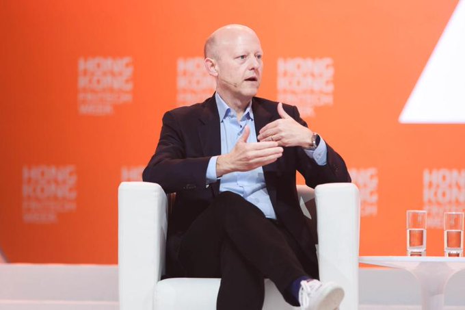 A bald man in a dark suit and light blue shirt sits in a white chair speaking at the 'HONG KONG FINTECH WEEK' event, gesturing with his hands.