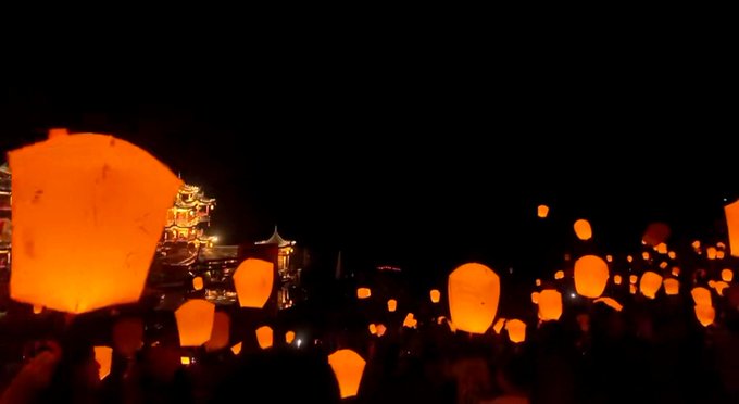 A nighttime festival scene features numerous glowing orange sky lanterns floating into the dark sky above a crowd, with a lit pagoda-style building in the background.