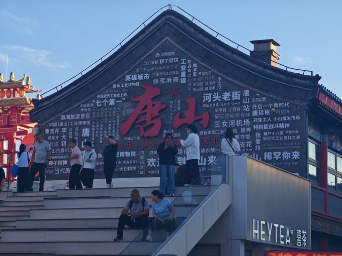 Visitors standing on a staircase in front of a large building with Chinese characters covering its facade, prominently featuring "唐山" (Tangshan) in red; a "HEYTEA 喜茶" sign is visible on the right.