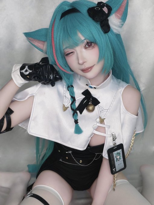 A cosplayer with teal hair and fox ears poses in a white and black costume with gold accents and a small bear hair accessory.