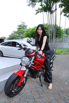 A woman with long dark hair, wearing a black top and leather pants, poses on a red Ducati motorcycle on a paved driveway.