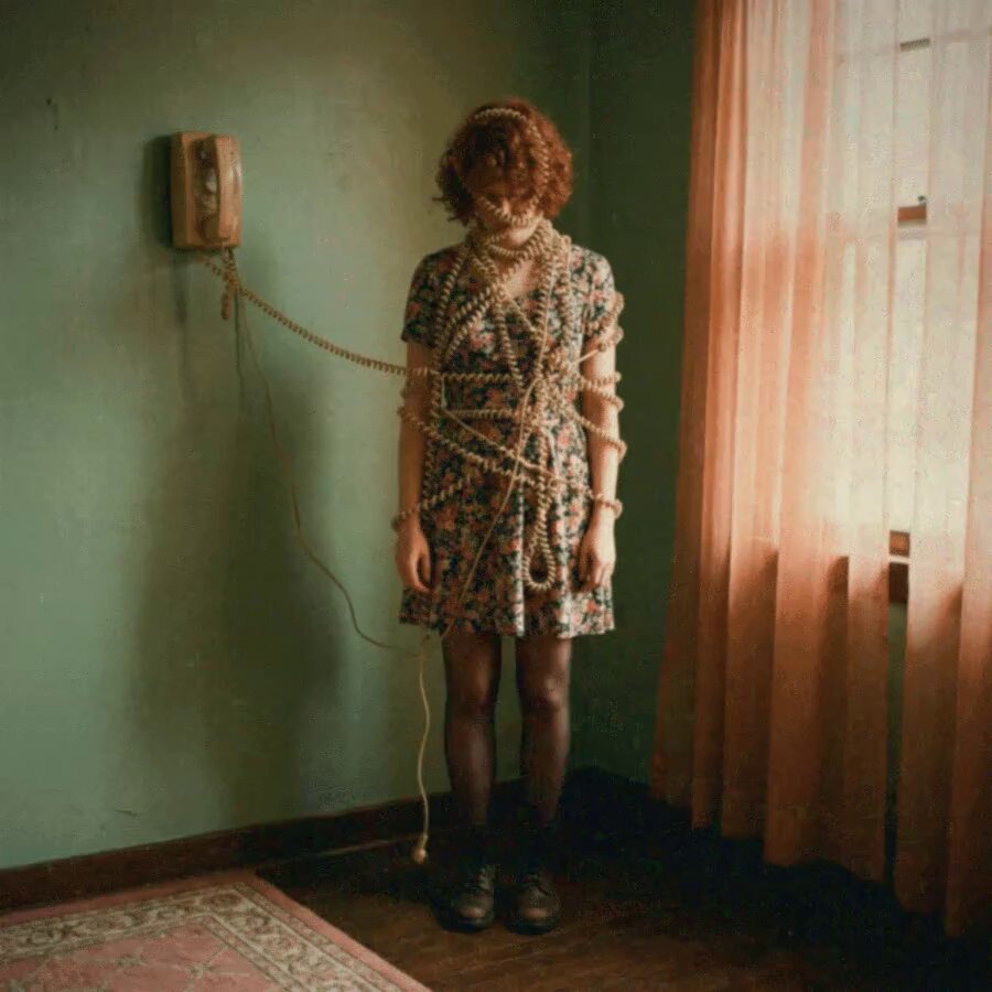 The image features a woman standing in front of a green wall, wearing a floral dress with tights and a headband. She has her hair tied up in a ponytail and is holding onto a cord that is connected to an old phone on the wall. The caption for this image reads "happy #GIFARTDAY, fam!: my new gif on @objktcom : 🜂boulali🜂 1/22 : 11 xtz".