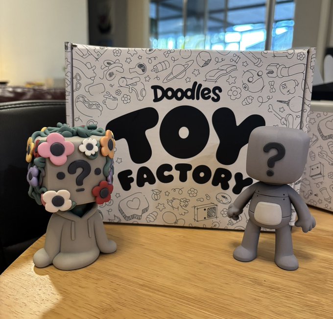 Two gray vinyl designer toys (one with a floral headdress, one plain with a question mark) displayed in front of a 'Doodles Toy Factory' box.