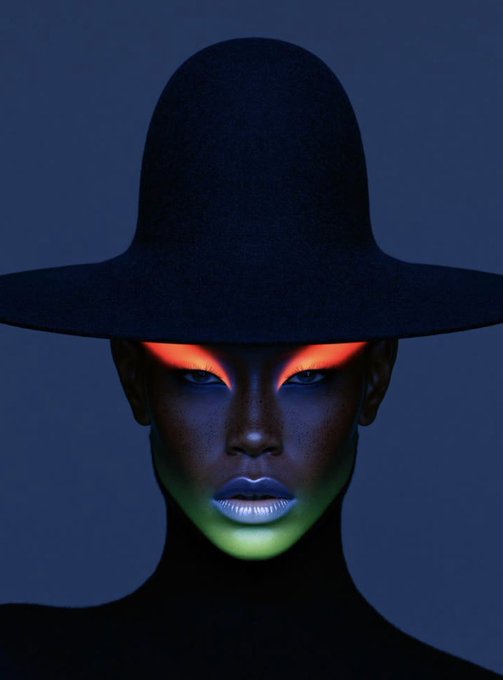 A dark portrait of a person wearing a wide-brimmed black hat, with glowing red and green light illuminating their face against a dark blue background.