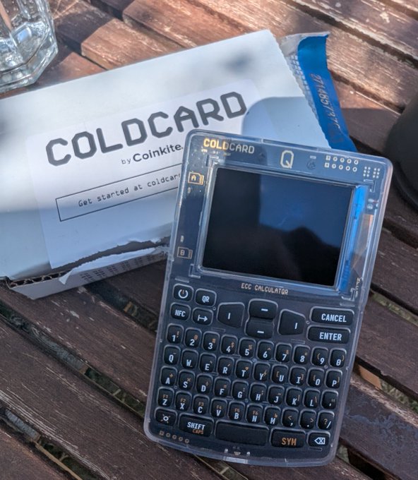 A transparent COLDCARD Q hardware wallet by Coinkite, featuring a screen and QWERTY keyboard, resting on its branded packaging on a wooden table.