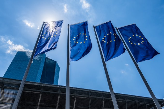 Four European Union flags wave in the foreground against a bright blue sky, with a modern glass skyscraper visible in the background—symbolizing the broader eurozone commerce landscape where only 0.2% of companies selling online accept cryptocurrency, according to a recent ECB survey.