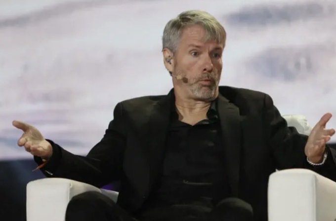 A grey-haired man in a black suit sits in a white chair on a stage, gesturing with both hands while speaking into a headset microphone.