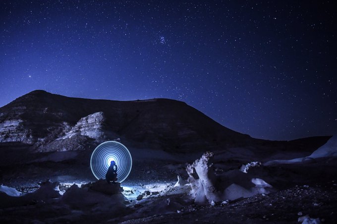 A person sits on rocky terrain under a starry night sky, illuminated by a glowing circular light painting behind them, with mountains in the background.