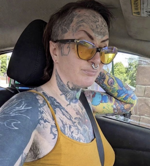 A heavily tattooed person with yellow-tinted sunglasses, a septum piercing, and a nose ring sits in a car, with an airbag warning label visible on the sun visor. The caption about body odor contrasts humorously with the bright yellow shirt and sunny expression, as nothing in the image suggests an unpleasant smell.