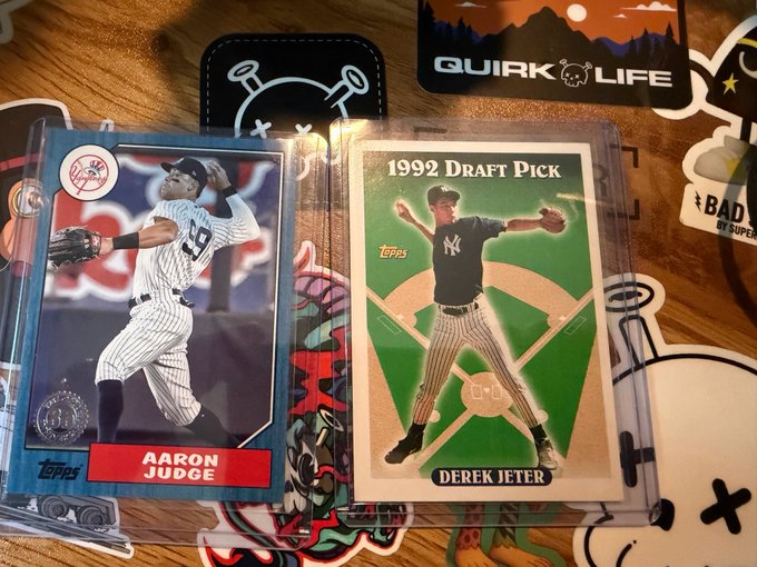 Two Topps baseball cards displayed on a wooden surface surrounded by stickers: an Aaron Judge Yankees card on the left and a 1992 Draft Pick Derek Jeter card on the right.