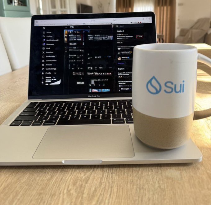 A MacBook Pro on a wooden desk displaying Twitter/X with a white and beige ceramic mug featuring the 'Sui' cryptocurrency logo in blue beside it.