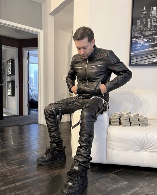 A man wearing a black leather jacket, leather pants with straps, and black boots sits on a white couch next to a stack of cash.