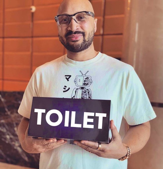 A bald man with glasses and a beard wearing a graphic t-shirt, holding a black sign that reads 'TOILET' in white letters.