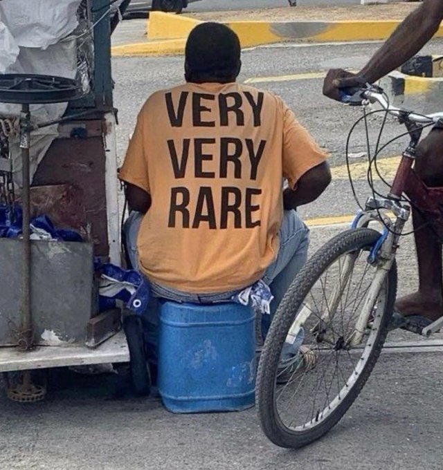 A man sits on a blue barrel on a street, wearing an orange t-shirt with bold black text reading 'VERY VERY RARE' on the back, next to a bicycle.