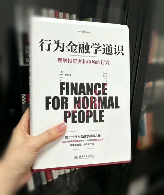 A hand holds the Chinese edition of "Finance for Normal People" by Meir Statman, with the cover featuring both Chinese characters "行为金融学通识" (subtitle "理解投资者和市场的行为") and the bold English title. The book aligns with the post's theme of behavioral finance, exploring the psychological biases like overconfidence and loss aversion that influence investor decisions in secondary markets.