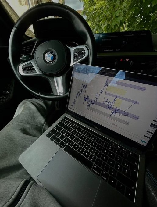 A laptop displaying a financial trading chart rests on a person's lap in the driver's seat of a BMW, with the M Sport steering wheel visible in the foreground. The scene humorously illustrates a "breakfast" focused on crypto trading rather than a meal, suggesting the trader's morning routine involves monitoring charts from their car within a $50 budget.