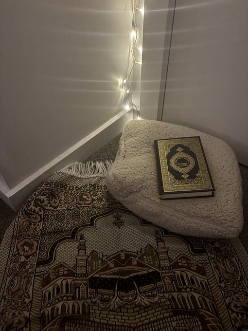 A cozy prayer corner setup with an Islamic Quran resting on a white cushion atop an ornate prayer rug depicting the Kaaba, with string lights on the wall behind.